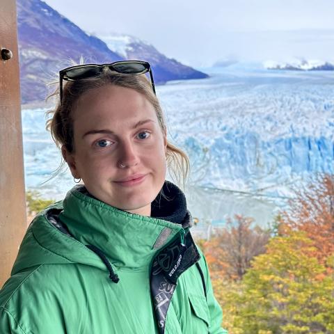 A girl wearing a green jacket stands outdoors with a glacier and mountains in the background.